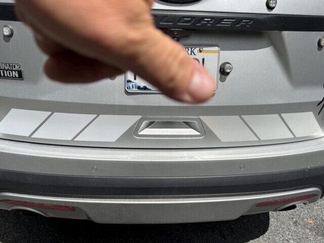 Rear bumper of a silver Ford Explorer with gray reflective shadow chevron tape applied across the bumper panel, shown in daylight.