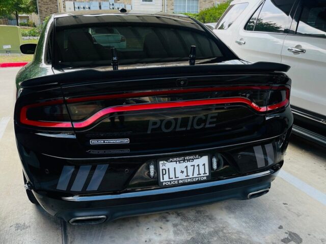 Black Dodge Charger Police Interceptor rear view showing gray reflective shadow chevron stripes on the bumper, parked in a residential driveway.