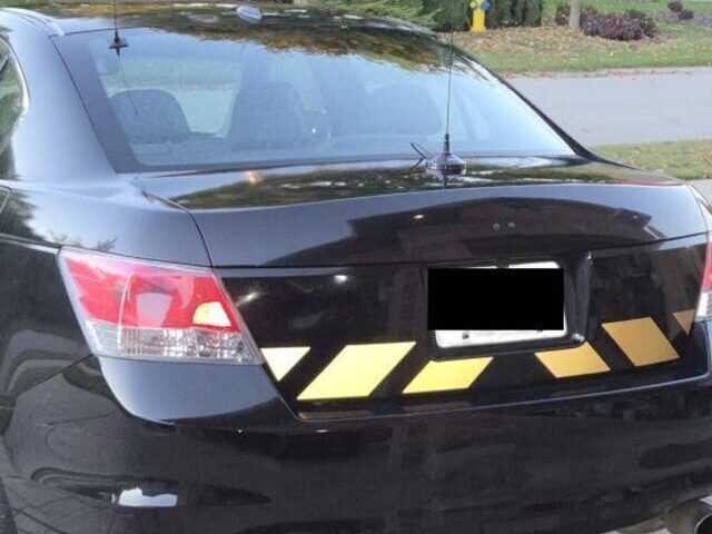 Black sedan rear view with yellow and gold reflective chevron stripes applied across the trunk area, photographed in a residential neighborhood during autumn.