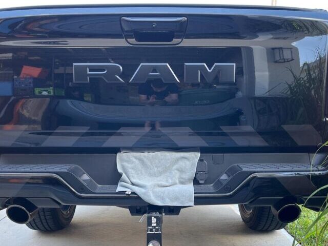 Black RAM pickup truck rear view showing dark gray reflective shadow chevron stripes on the tailgate, photographed in a garage driveway during daylight.