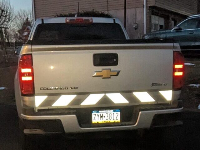 Silver Chevrolet Colorado pickup truck rear view at dusk with white reflective chevron tape glowing brightly on the bumper, headlights illuminating the reflective material.