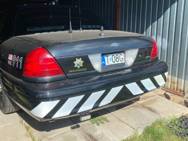 Black Ford Crown Victoria Police Interceptor with bold white reflective chevron stripes spanning the full width of the rear bumper, parked under a metal carport.